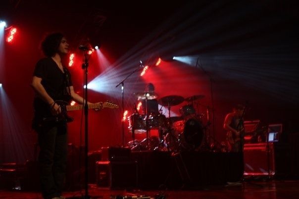Live concert with guitarist and drummer on a red-lit stage, bright spotlights behind them
