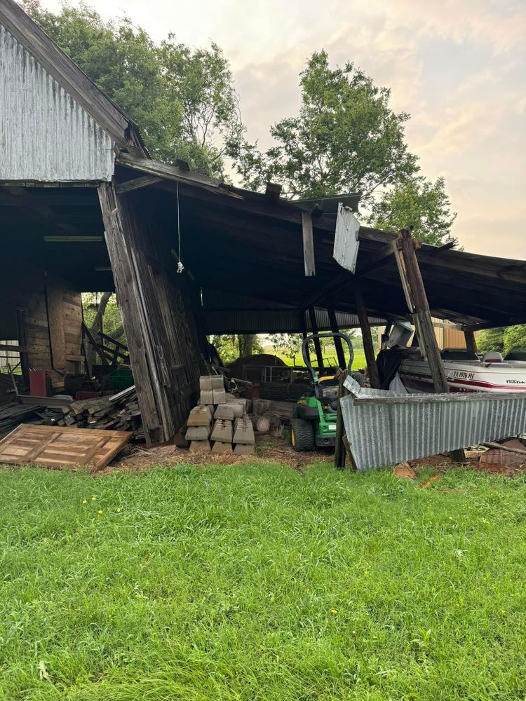 A barn with a broken roof is sitting in the middle of a grassy field.