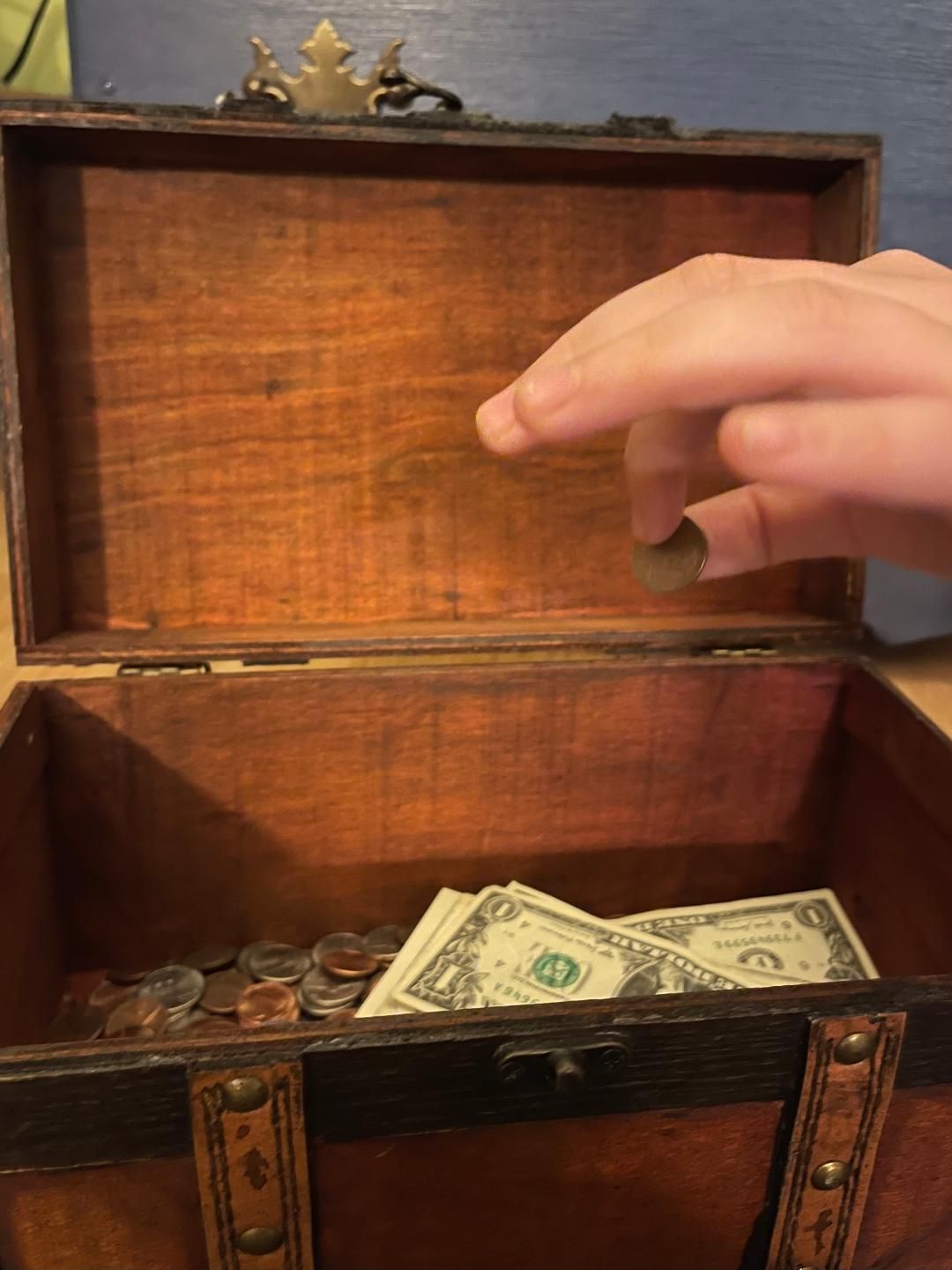 Boy's fingers dropping a penny into a wooden treasure chest bank with coins and paper bills in the b