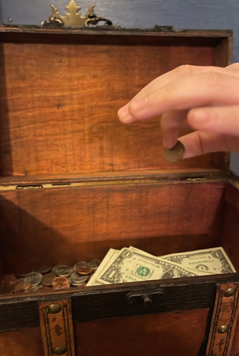 Boy's fingers dropping a penny into a wooden treasure chest bank with coins and paper bills in the bottom.