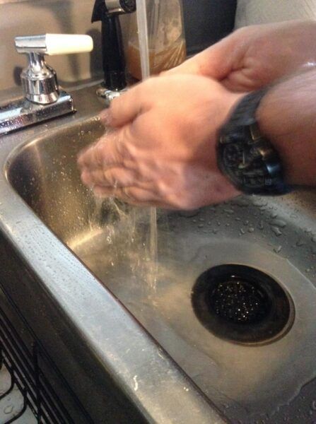 Man with large black watch washing his hands in water over a stainless steel sink