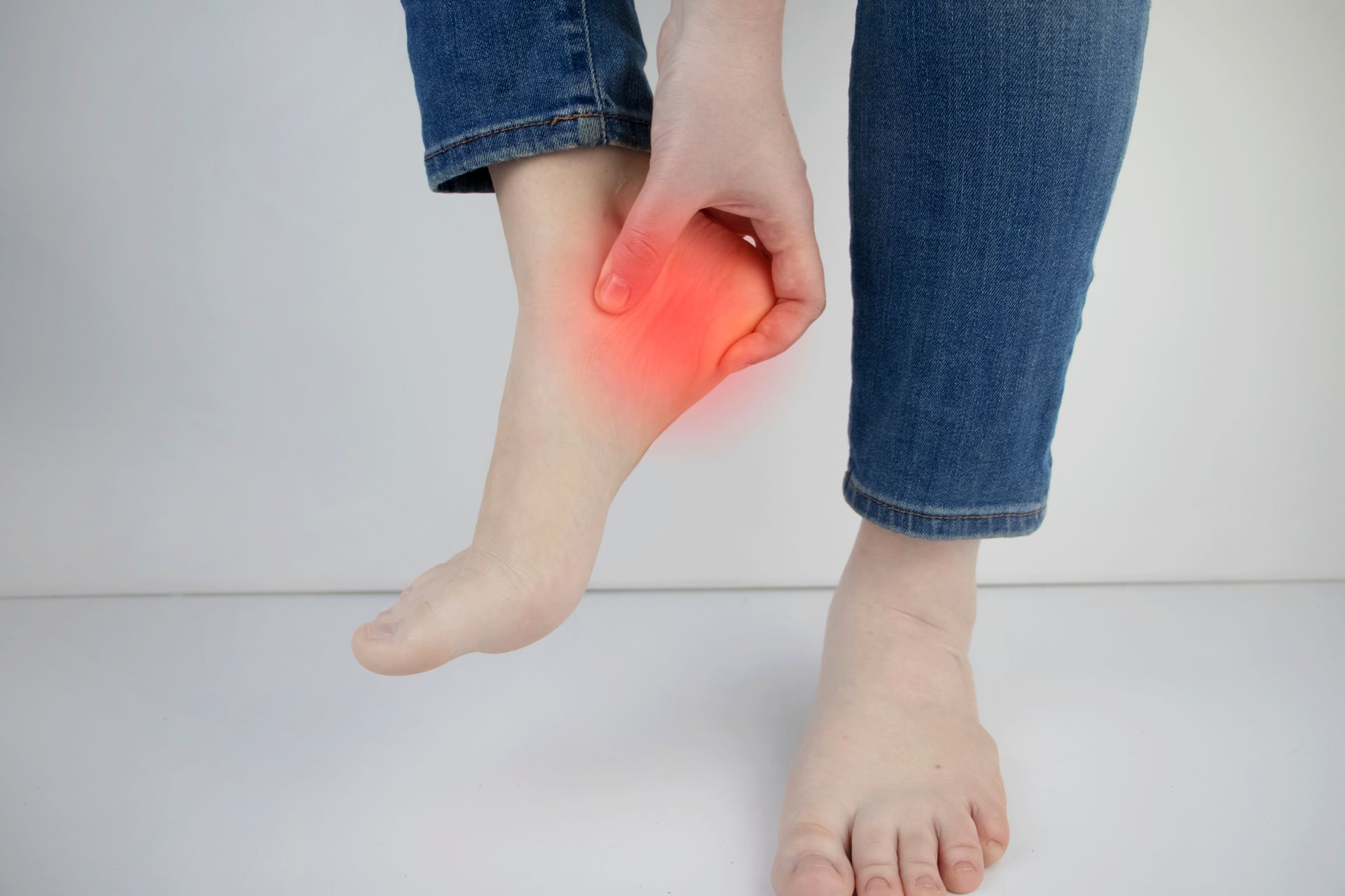 Person holding foot, red highlight on heel, indicating pain. Wearing jeans, neutral background.