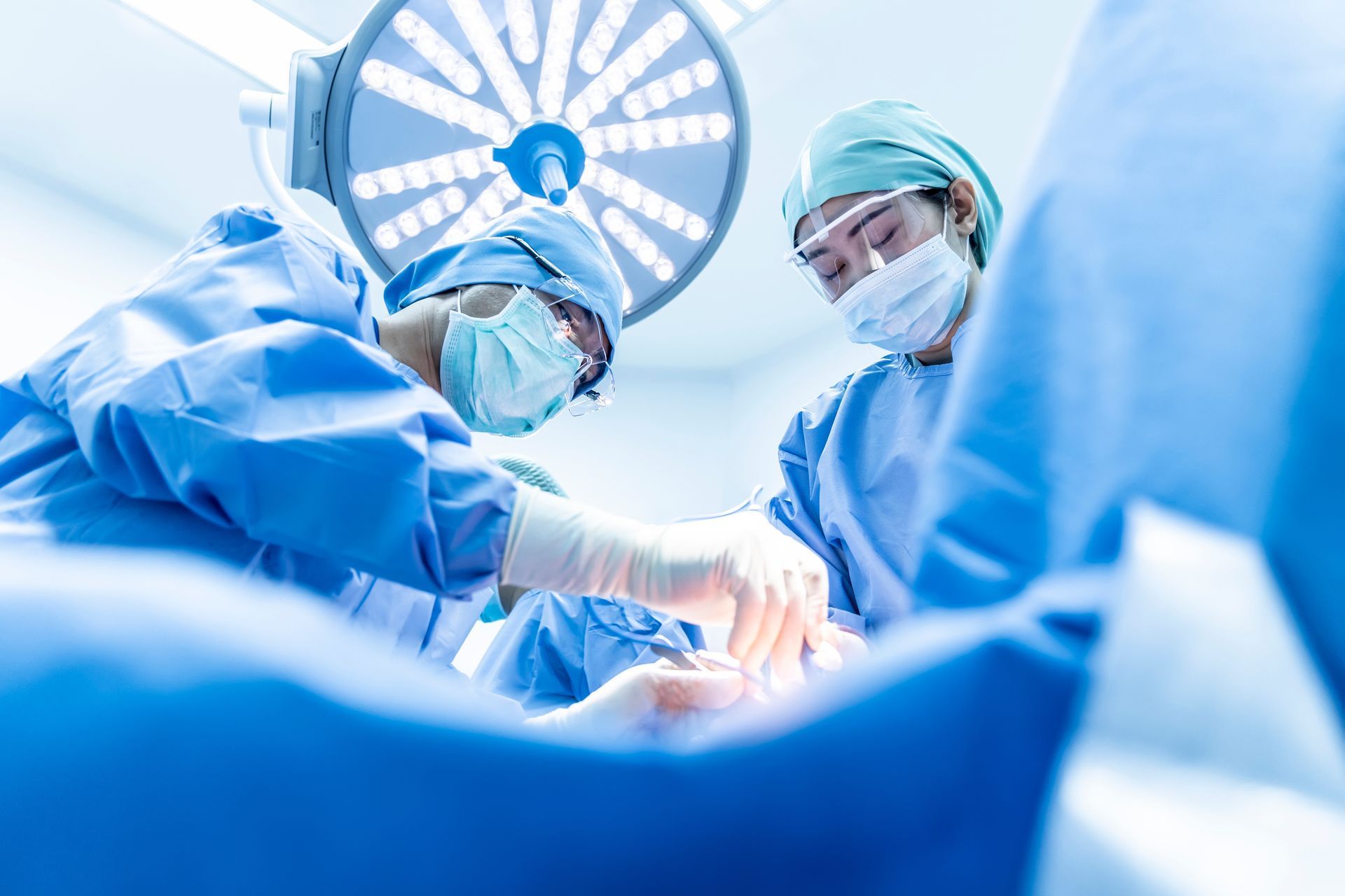 Surgeons in blue scrubs operate under bright lights in an operating room.