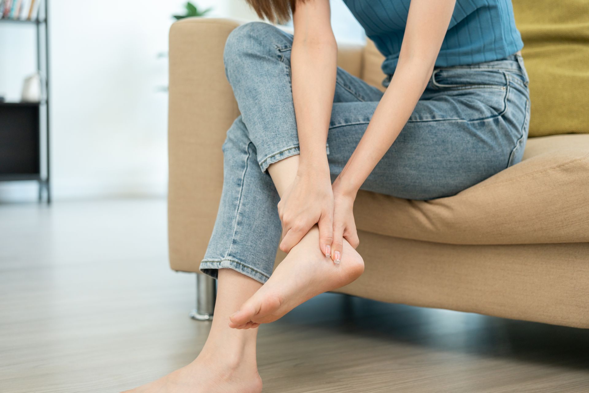 Woman sitting on a beige sofa, holding her foot. She's wearing jeans and a blue top.