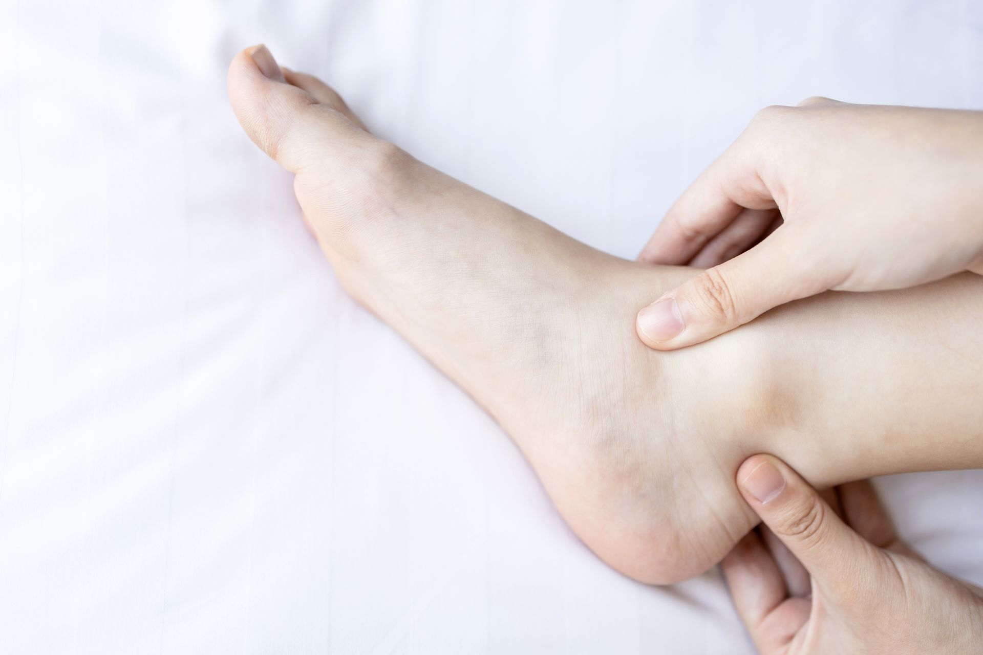 Person's hands gently touching and examining their ankle and foot resting on white fabric.