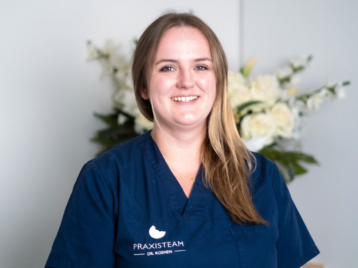 Eine Frau in einem blauen Kittel lächelt vor einer Vase mit Blumen.