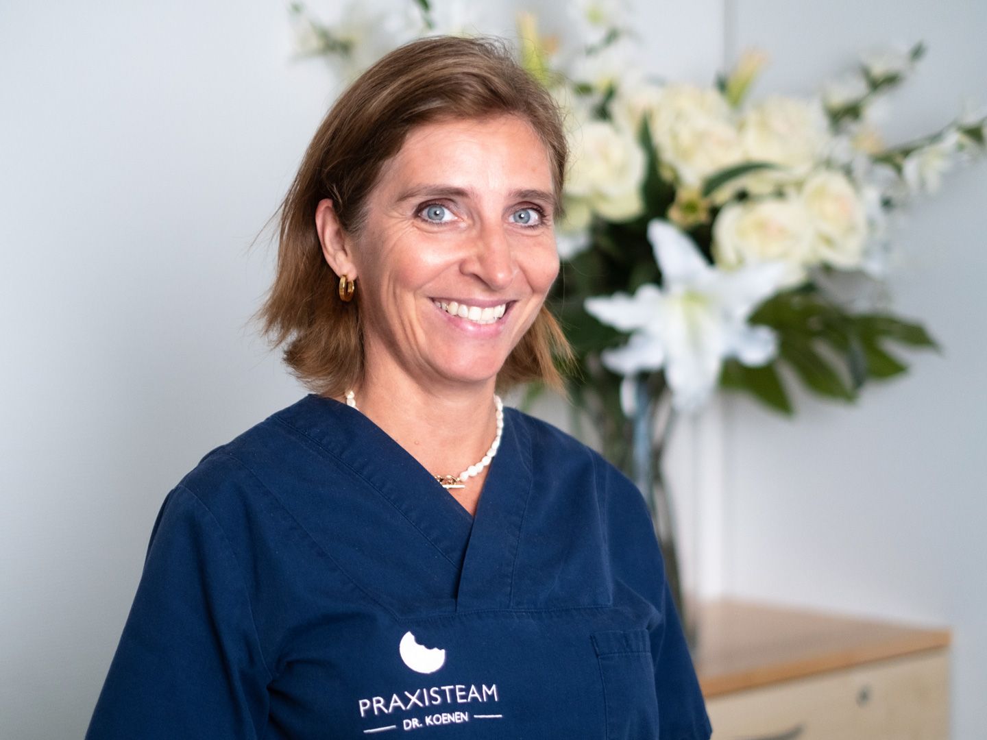 Eine Frau in einem blauen Kittel lächelt vor einer Vase mit Blumen.