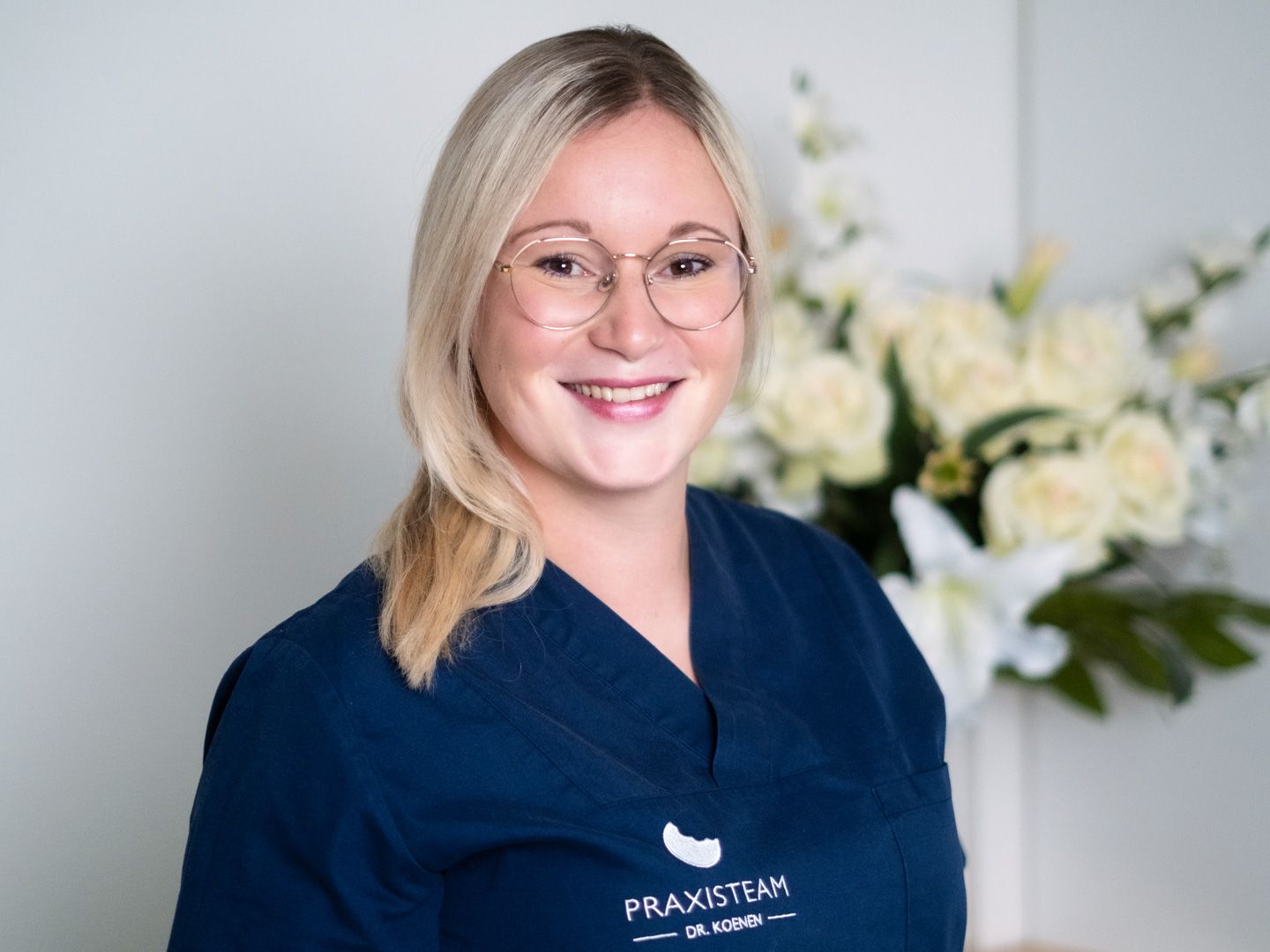 Eine Frau mit Brille und blauem Kasack lächelt vor einer Blumenvase.