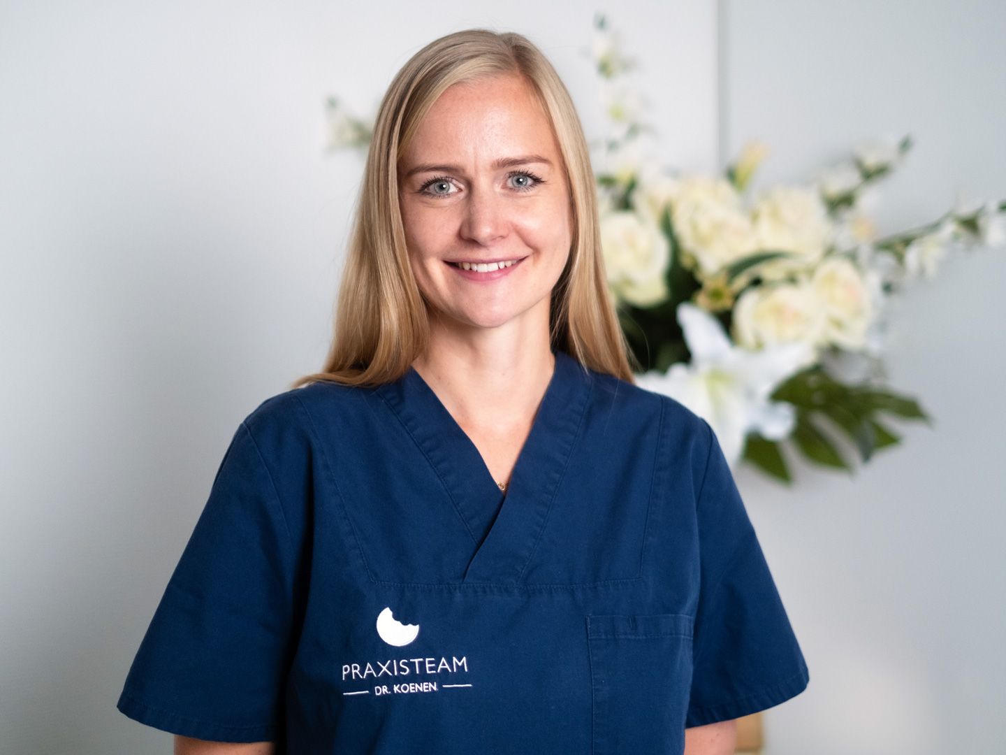 Eine Frau in einem blauen Kittel lächelt vor einem Blumenstrauß.