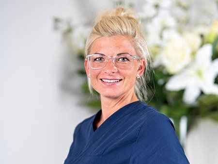 Eine Frau mit Brille und blauem Kittel lächelt vor einer Blumenvase.