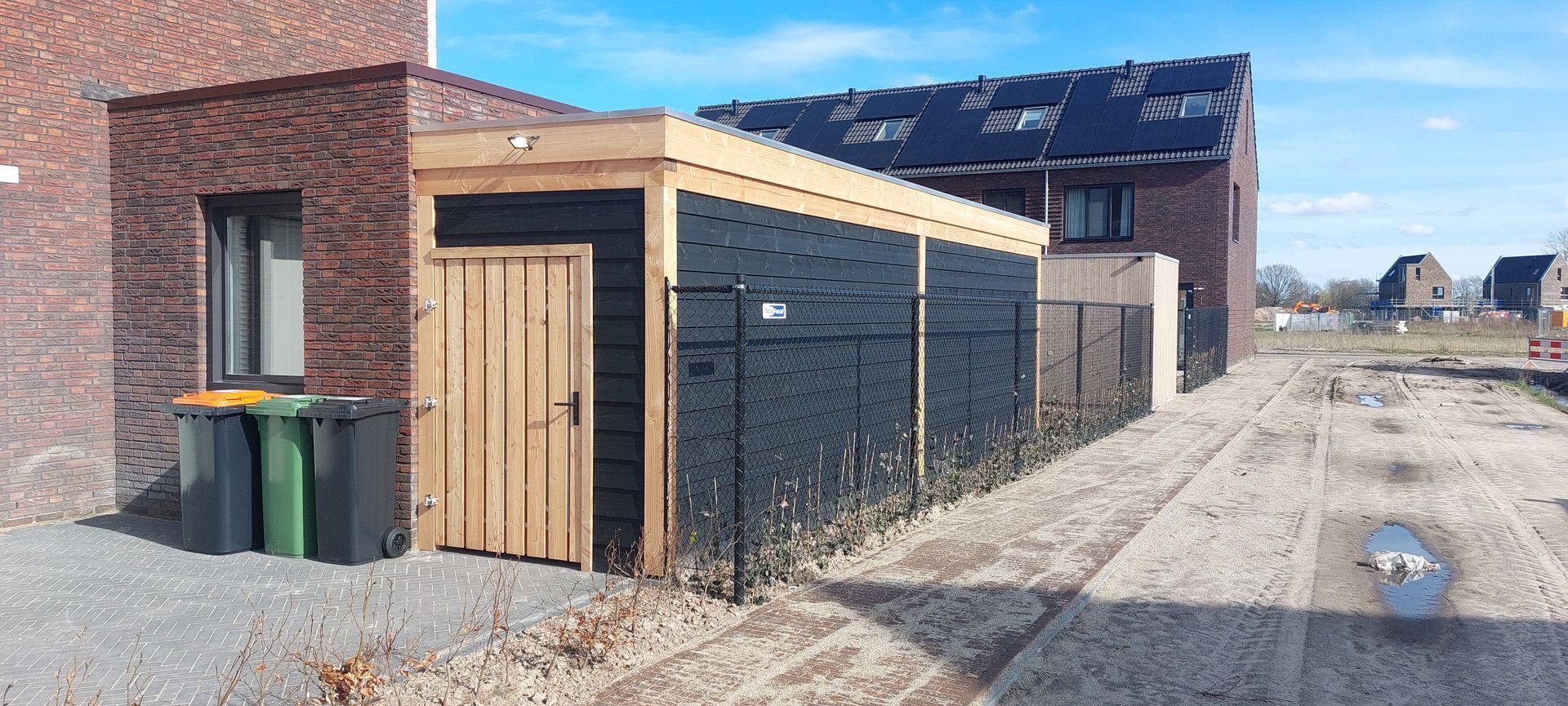 Een rij vuilnisbakken naast een bakstenen gebouw en een houten schuur op een grindpad.