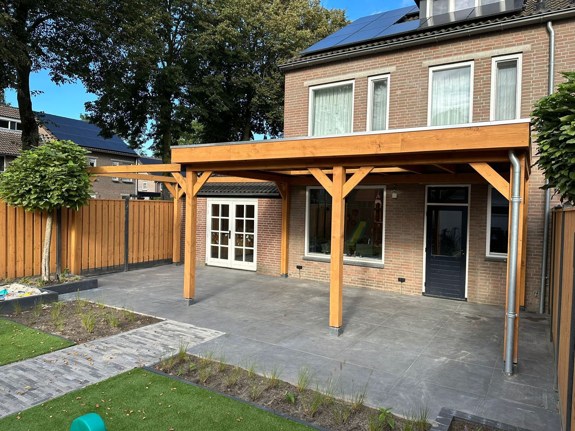 Een houten pergola, bevestigd aan een bakstenen huis, boven een betonnen terras. | Smans Houtbouw