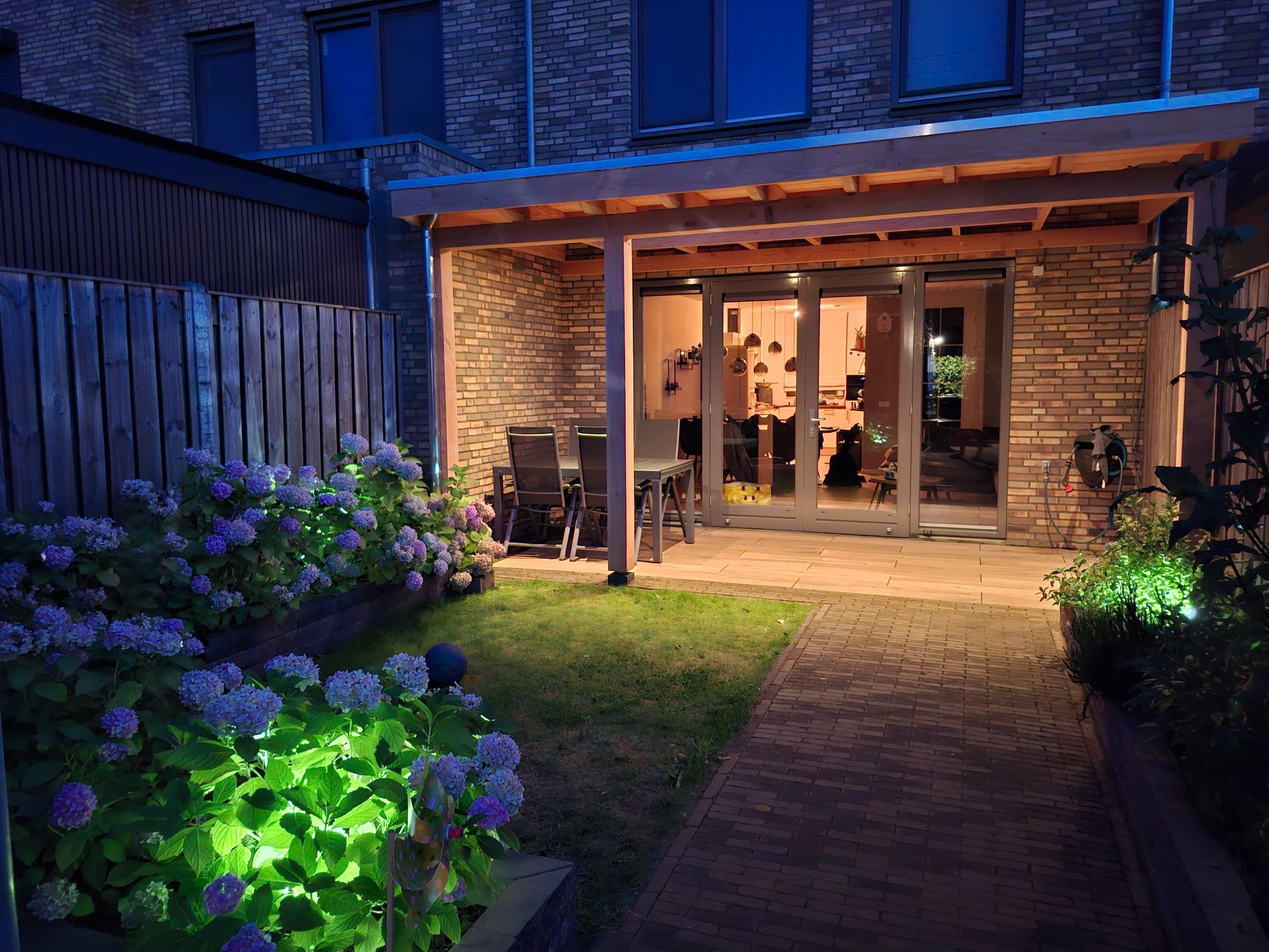 Nachtelijk uitzicht op een achtertuin met een verlicht terras en een tuin. Hortensia's bloeien op de voorgrond.