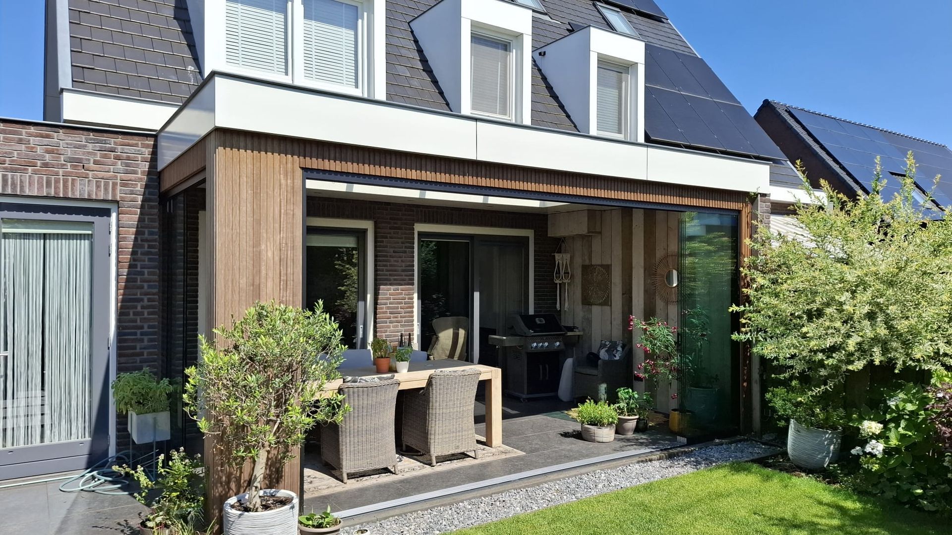 Terras met tafel en stoelen, grenzend aan een bakstenen huis met een donker dak en zonnepanelen.