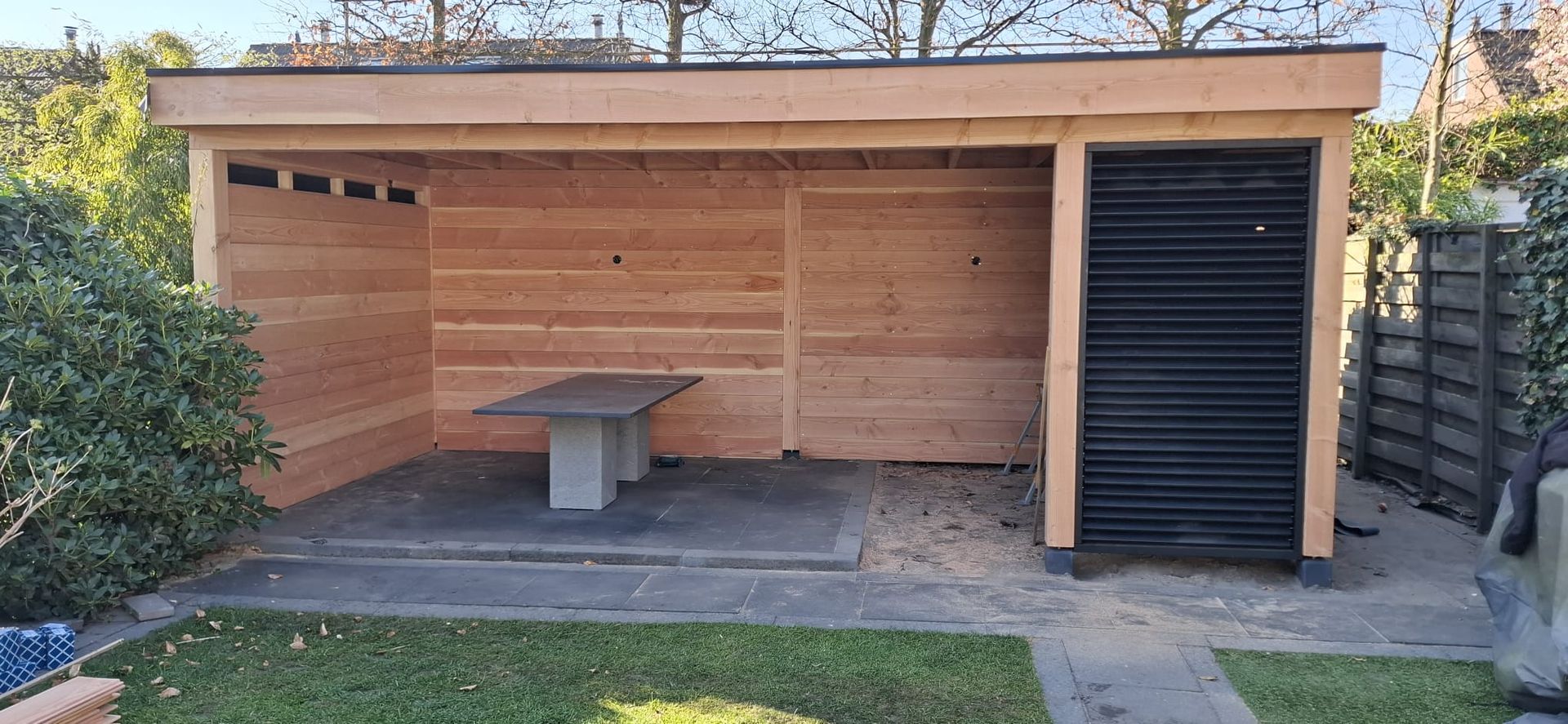 Een houten buitenconstructie met een tafel, een zwarte wand met luiken en een grasveld.