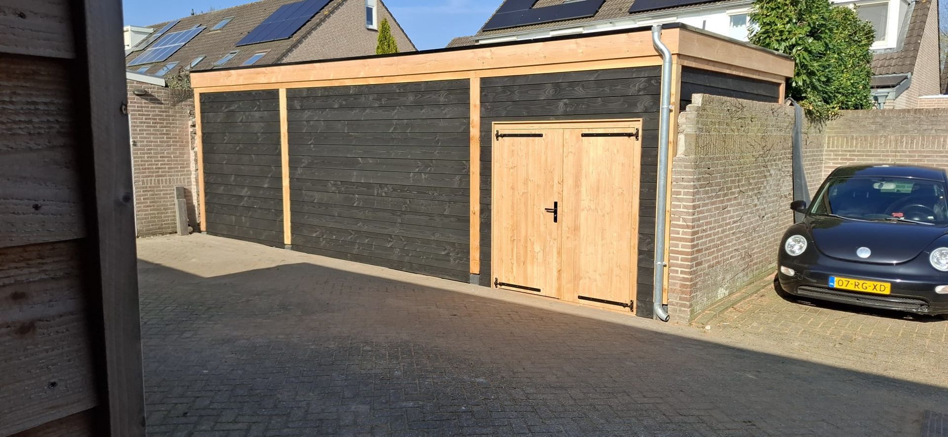 Een garage van zwart en licht hout met een auto ernaast geparkeerd.