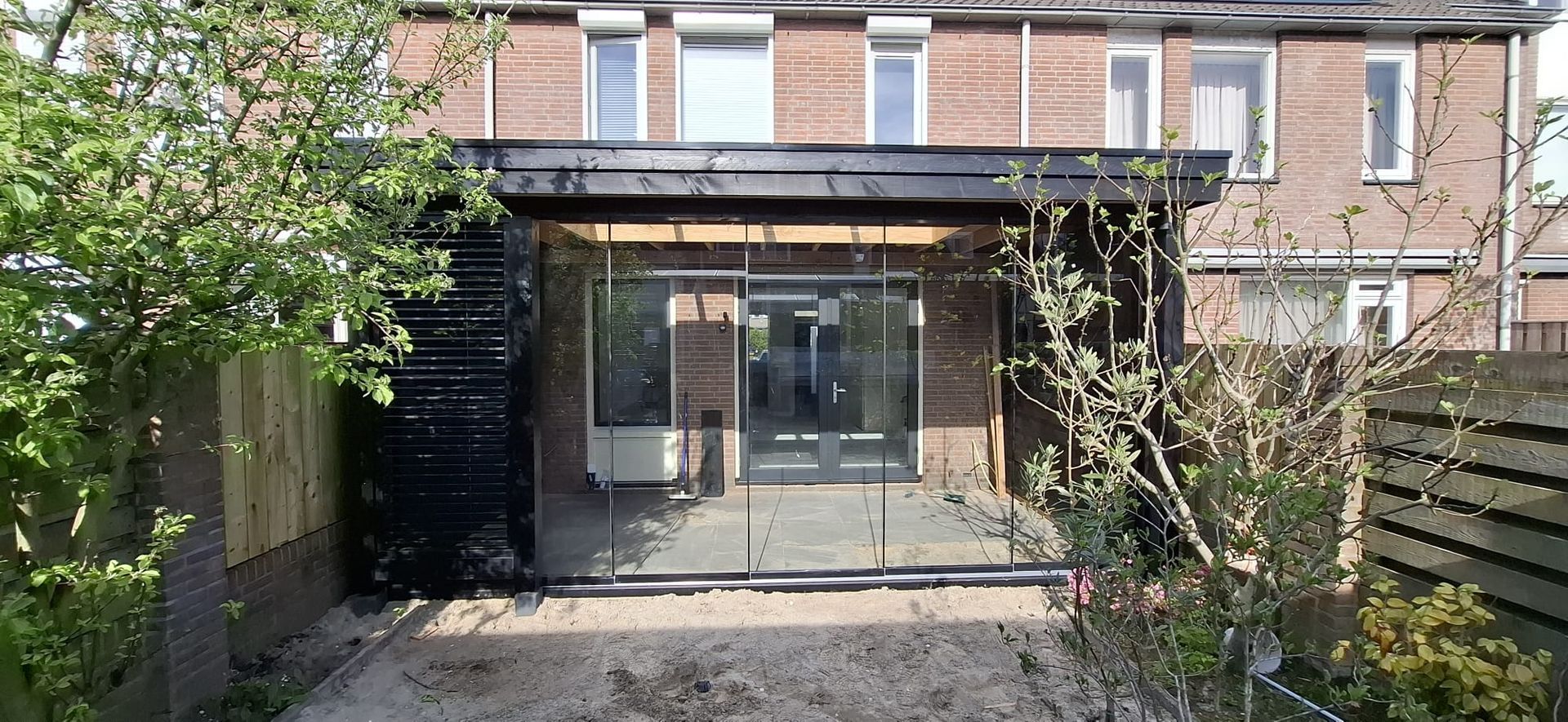 Een modern gebouw met een glazen voordeur en een zwart dak, omgeven door bomen en een gebouw van bruine baksteen.