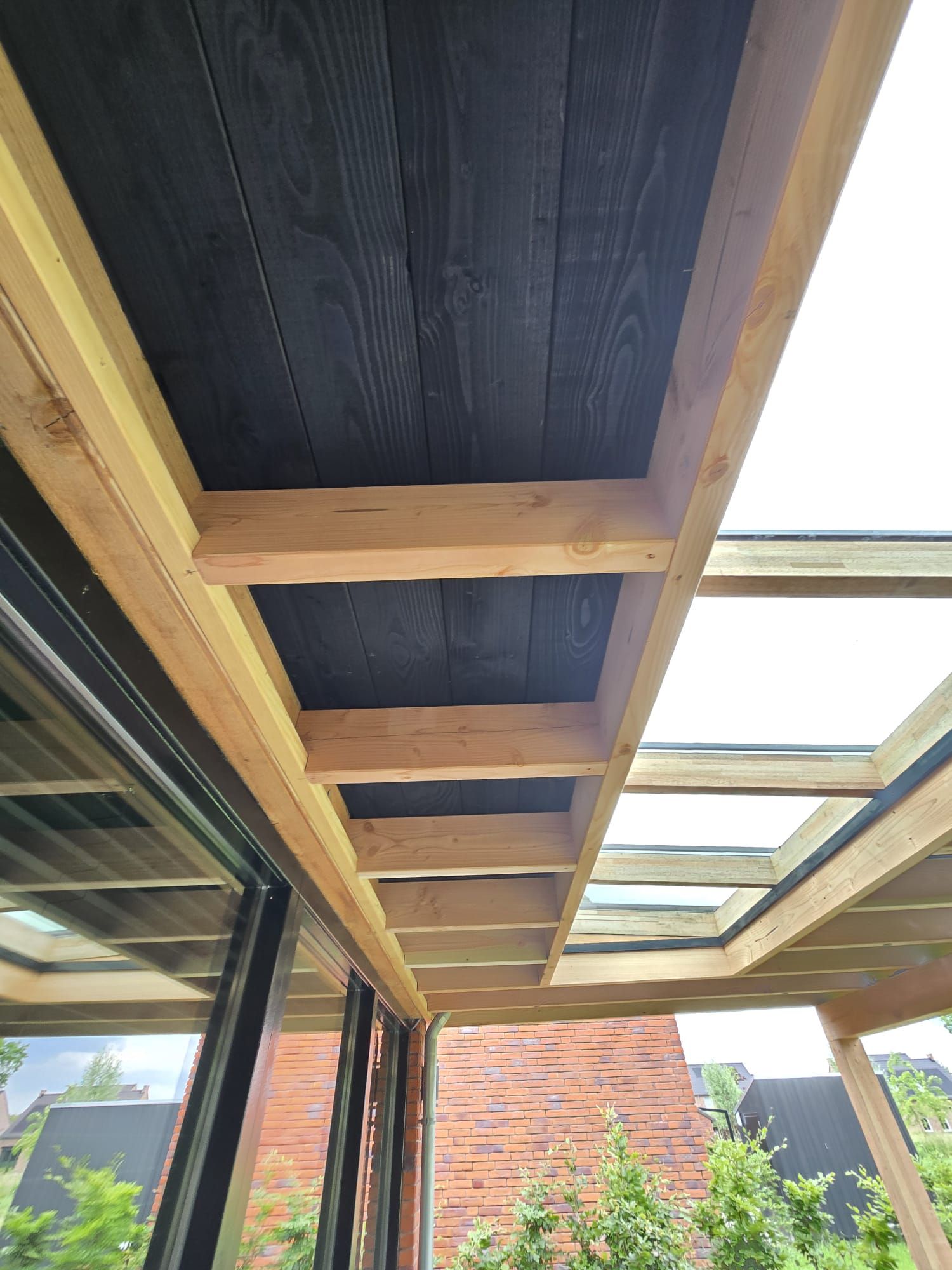 Houten pergola met zwarte planken en glazen panelen naast een gebouw.