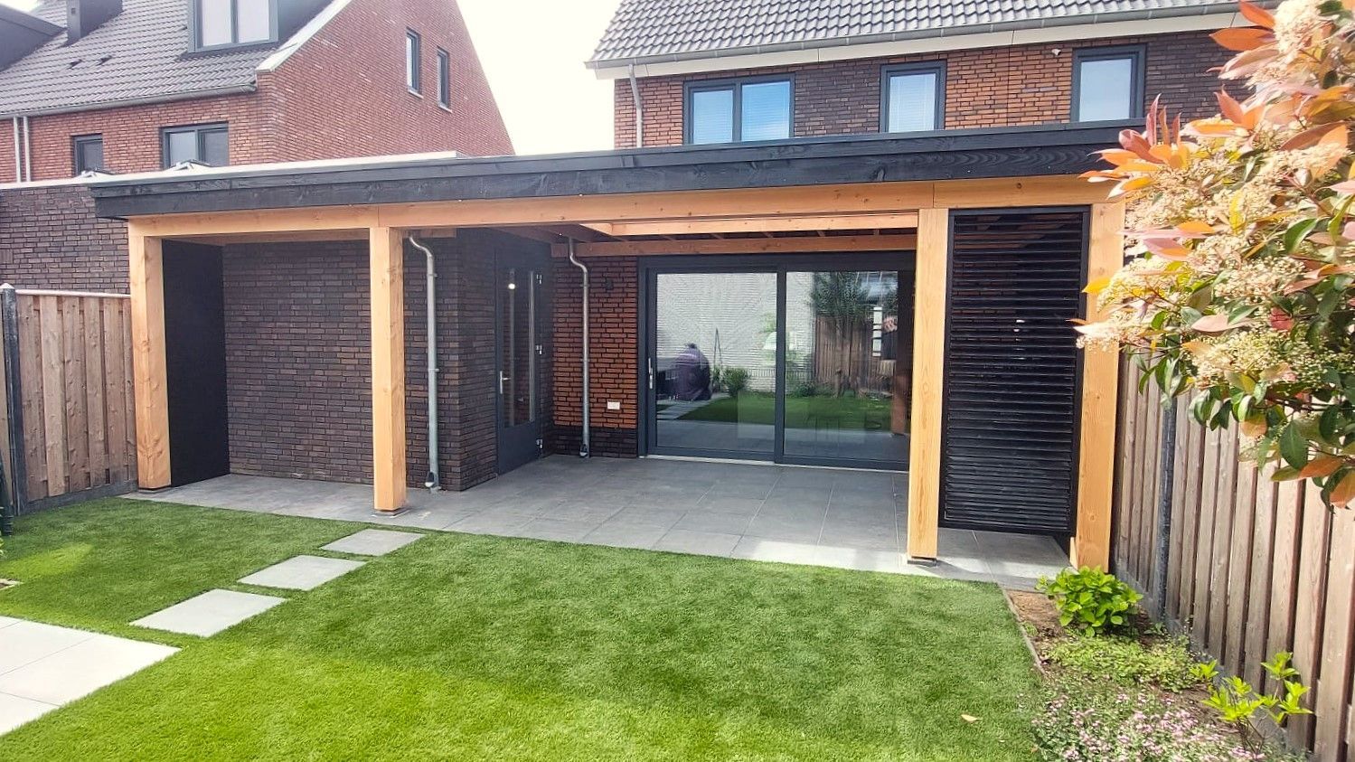 Carport met bakstenen muren en houten balken, met uitzicht op een grasveld met stapstenen.