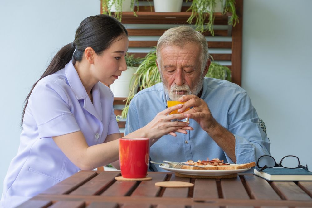 A nurse is feeding an elderly man a glass of orange juice.