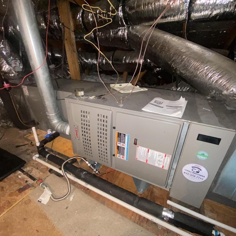 Furnace unit in an attic, with ductwork and exposed wires. Gray metal, black and silver pipes.