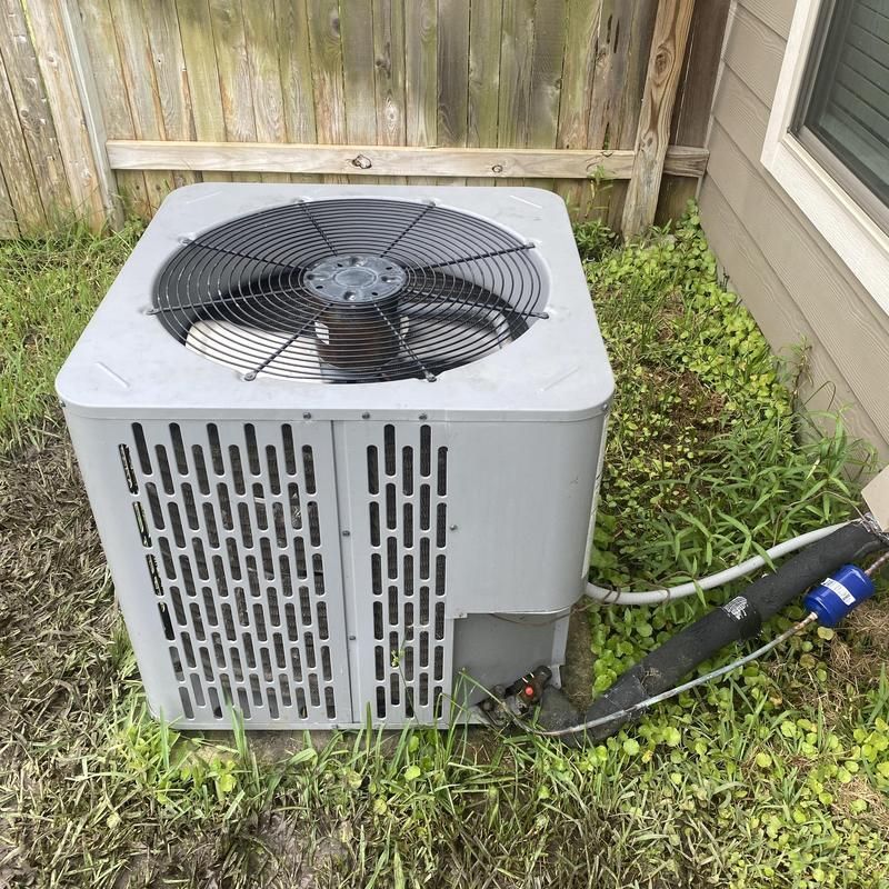 Air conditioning unit outside a building, surrounded by grass, near a wooden fence.