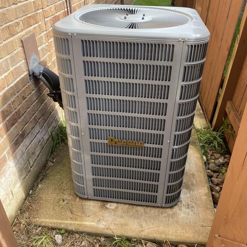 Air conditioning unit, grey and silver, outdoors next to a brick wall and wooden fence on concrete.