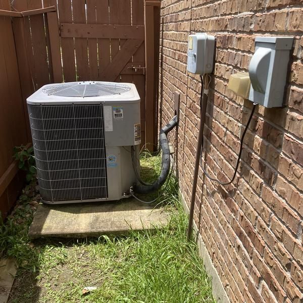 Air conditioning unit next to a brick wall, with electrical boxes and a wooden fence in the background.