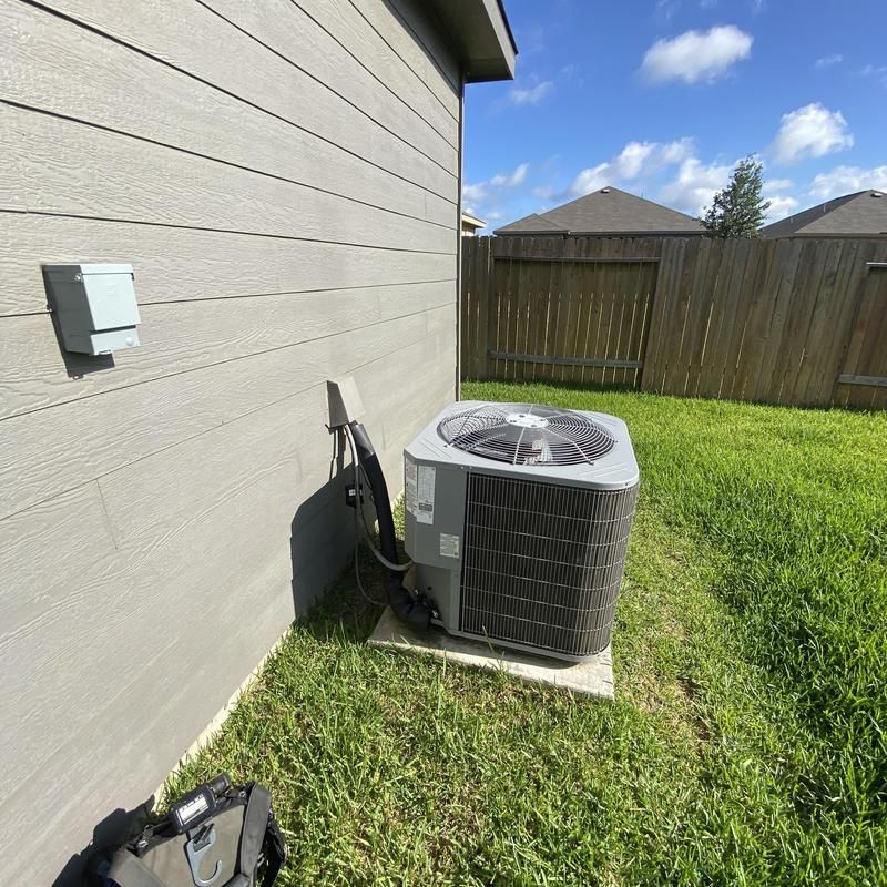 An outdoor air conditioning unit next to a house, with a power box on the wall and a fence in the background.
