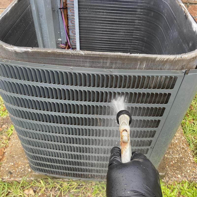 Person in black glove sprays an air conditioner unit with a water hose outside.