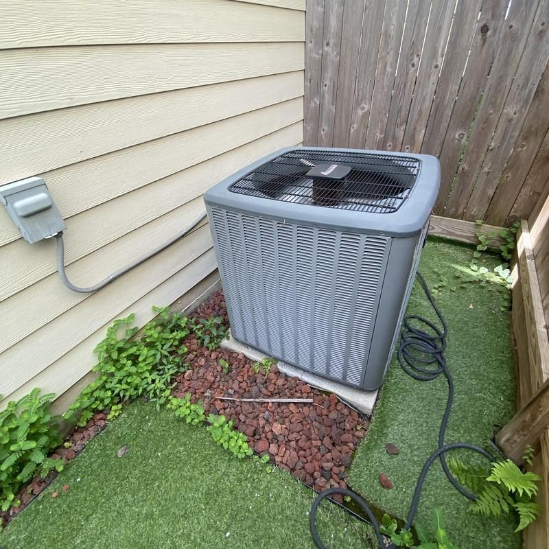 Gray air conditioning unit next to beige siding and a wooden fence, surrounded by plants and lava rock.