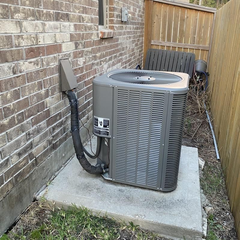 An air conditioning unit on a concrete pad next to a brick wall and wooden fence.