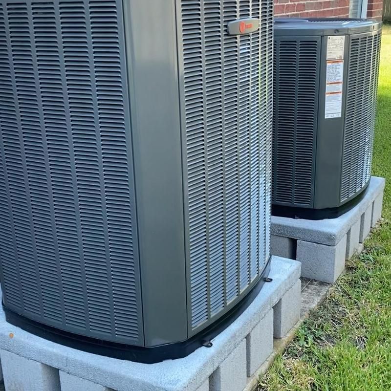 Two gray air conditioning units on concrete block bases in a yard.