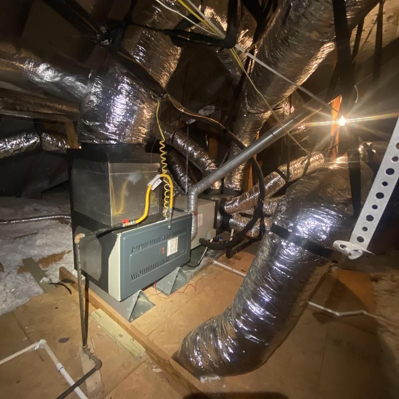 HVAC unit in an attic with silver ductwork and wiring. Bright light shines, illuminating the space.