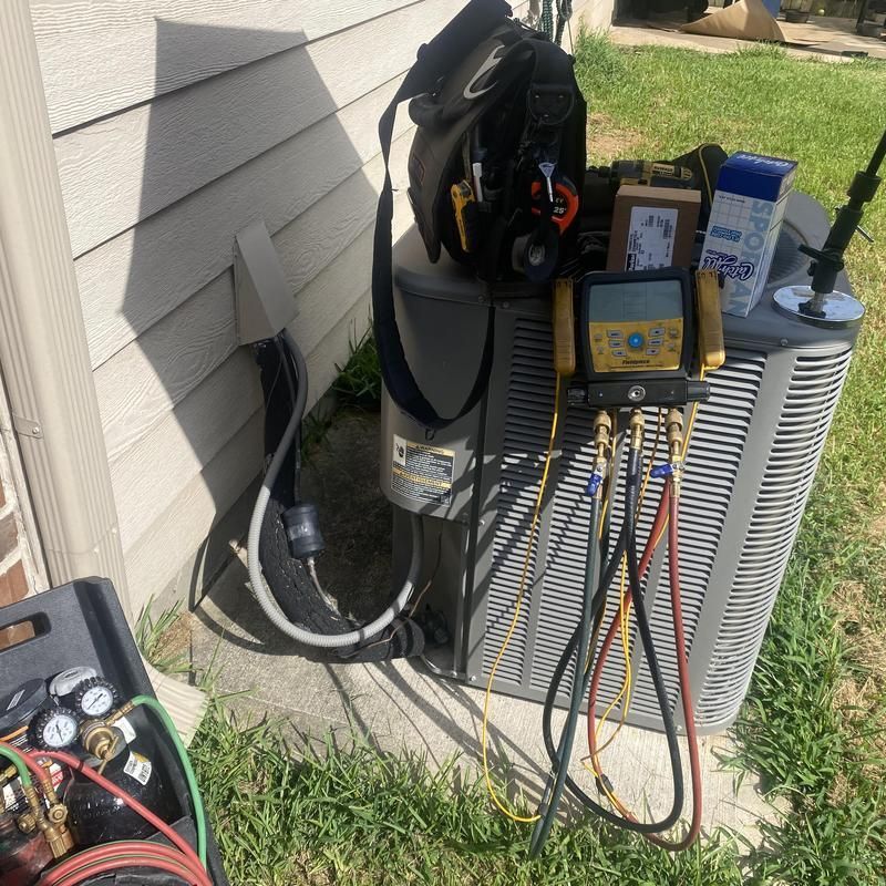 HVAC technician servicing an AC unit. Tools, gauges, and hoses visible. Outdoor setting, sunny day.