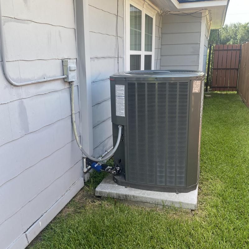 Air conditioning unit outside a building on a concrete base, connected to piping and electrical box.