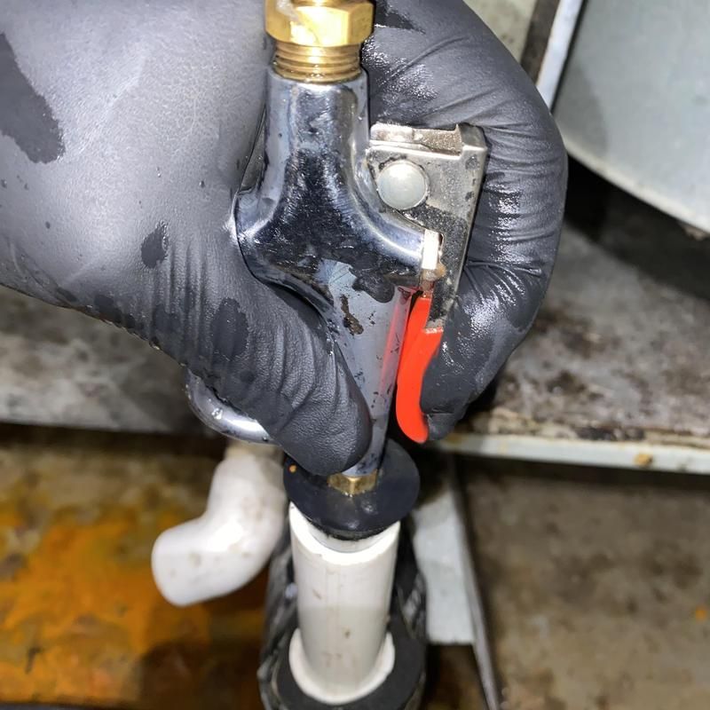 Gloved hand holding a metal nozzle connected to a white pipe, spraying water. Red trigger.