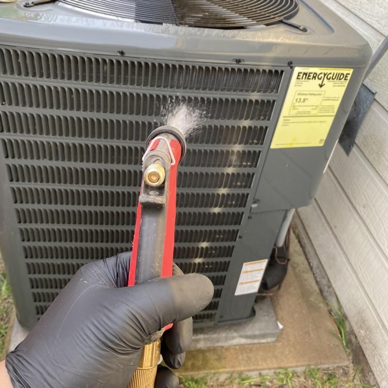 Gloved hand cleaning an AC unit's exterior fins with a spray nozzle, removing debris.