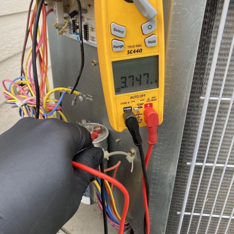 A gloved hand uses a multimeter to test electrical components on an AC unit.