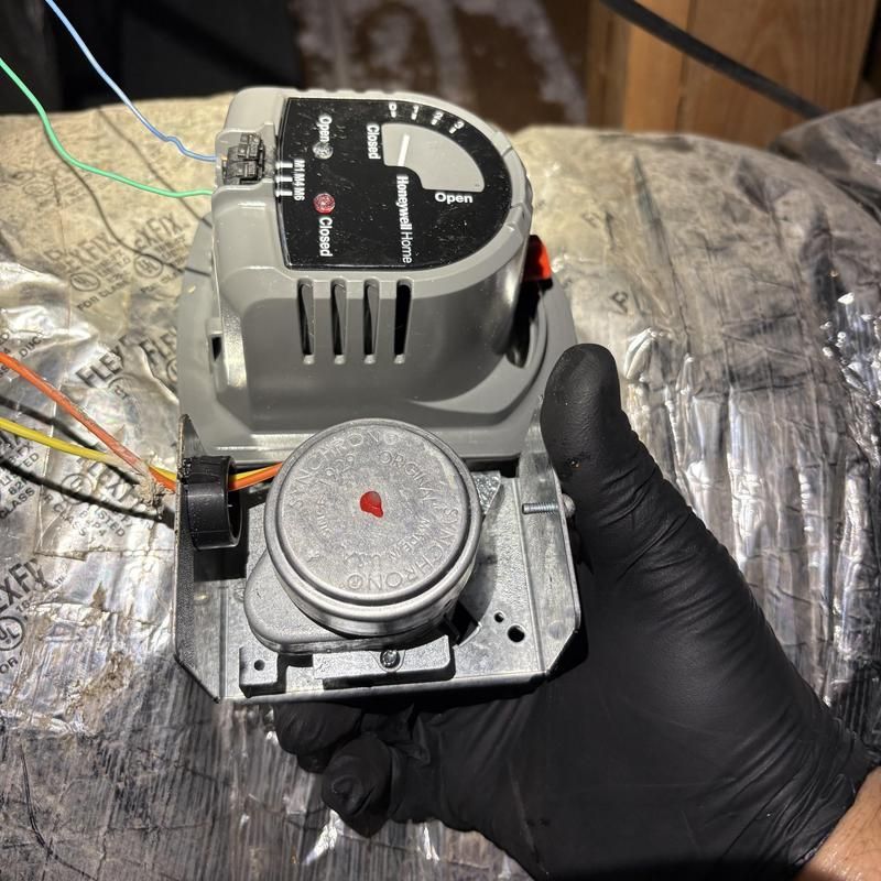 Black gloved hand holding HVAC actuator, with wires attached, installed in ductwork.