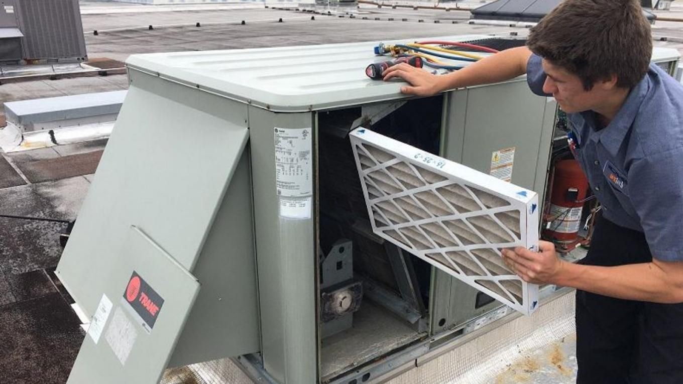 HVAC technician replacing an air filter in a rooftop unit. Grey unit, blue shirt, outdoor setting.