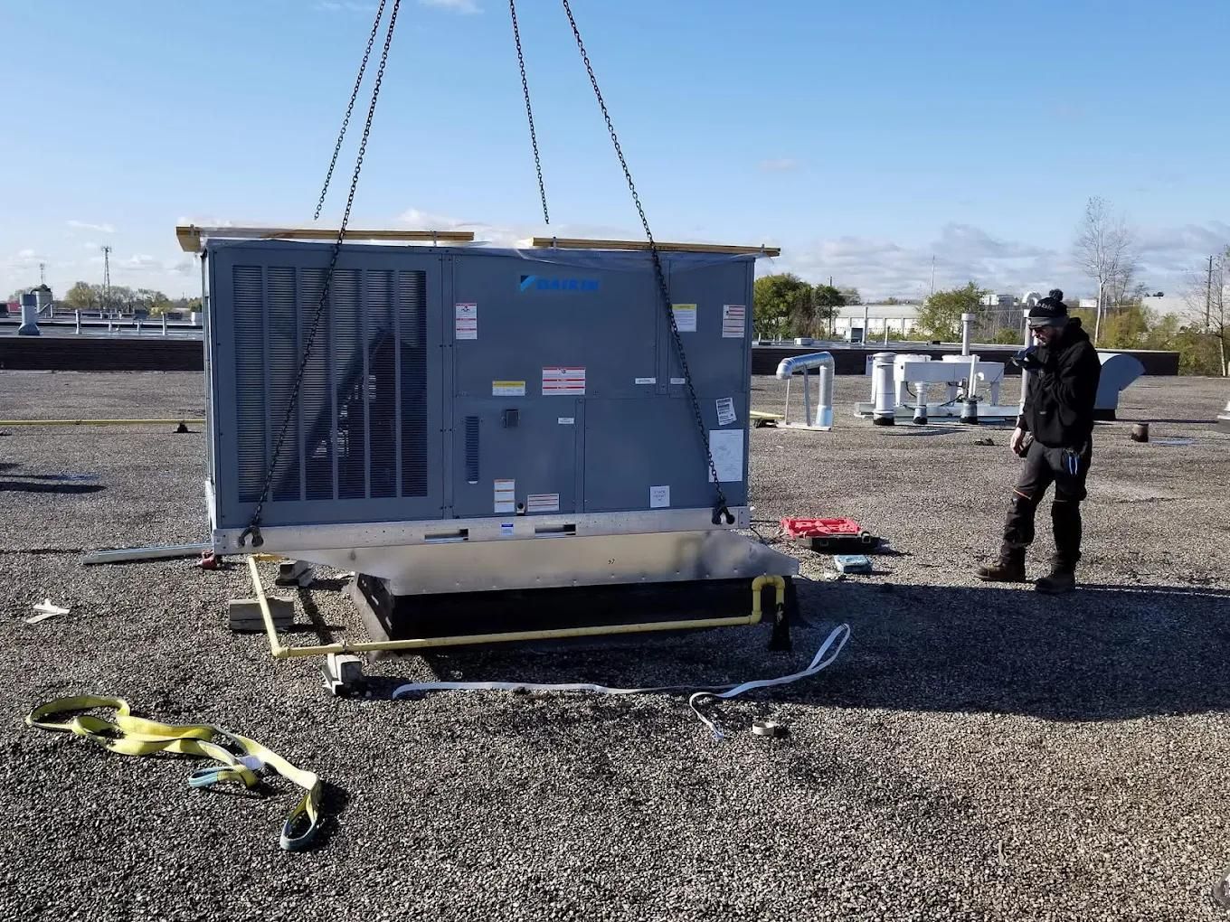 HVAC unit being lifted by chains on a rooftop with a worker observing.