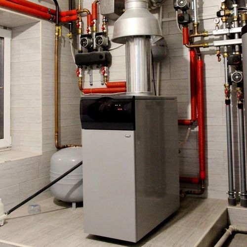 A gray boiler with red and copper pipes in a utility room, with a silver exhaust vent above.