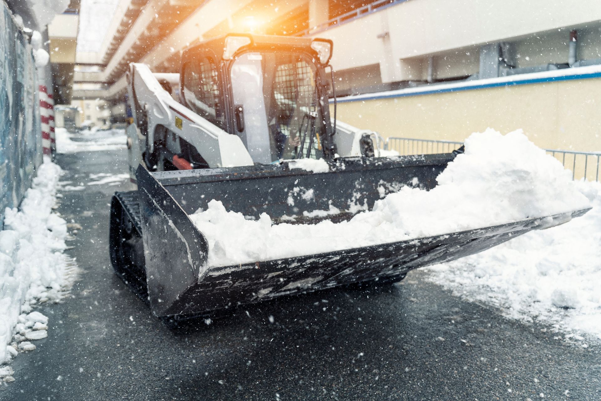 A skid steer with a snowplow clearing snow from a road during a snowfall.