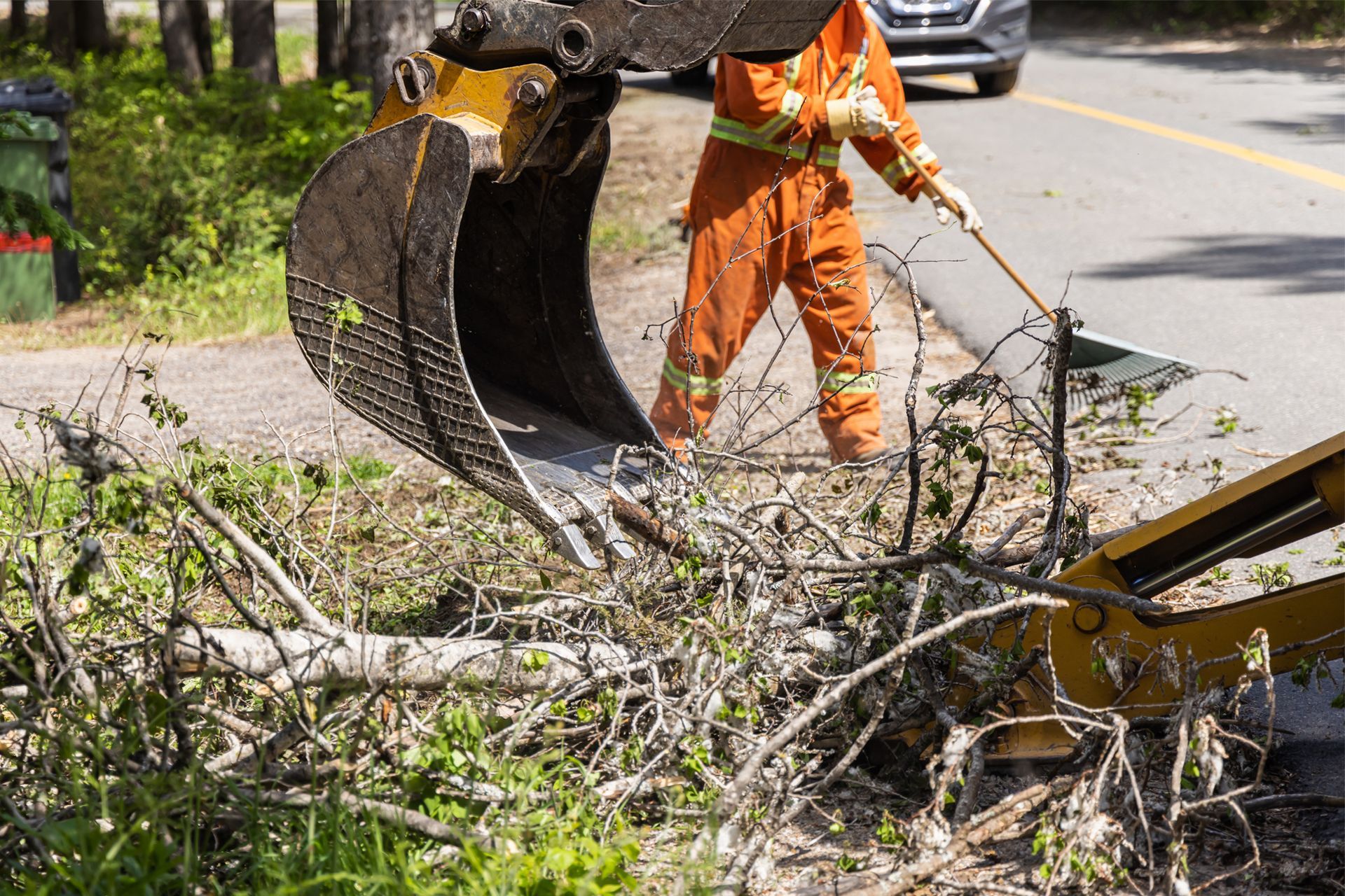 Worker in orange suit using excavator to clear brush alongside a road.
