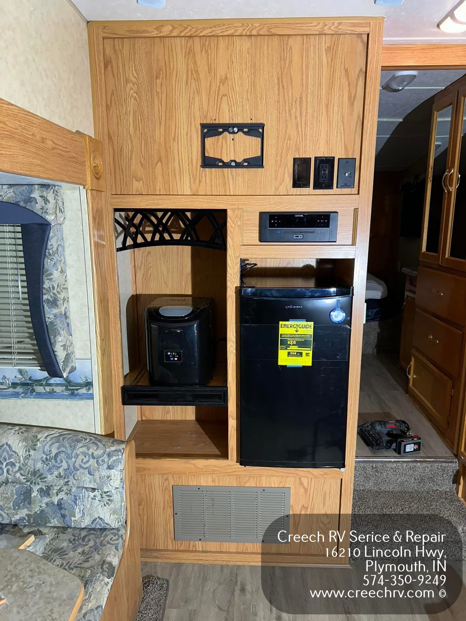 RV interior with wood paneling, featuring a small refrigerator, shelf, and TV mounting bracket.