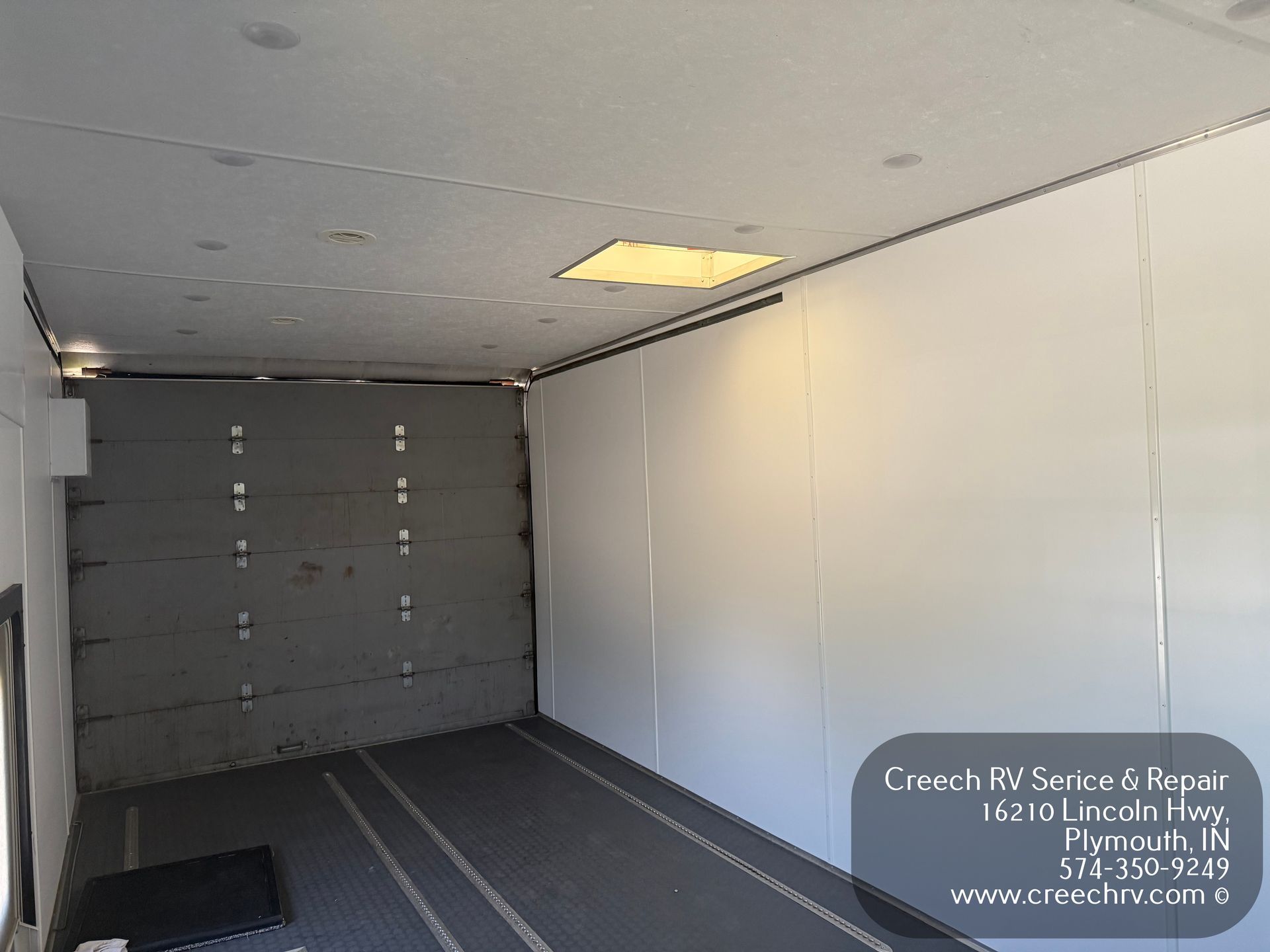 Interior view of an empty RV cargo area with white walls, black floor, and a closed gray rear door.