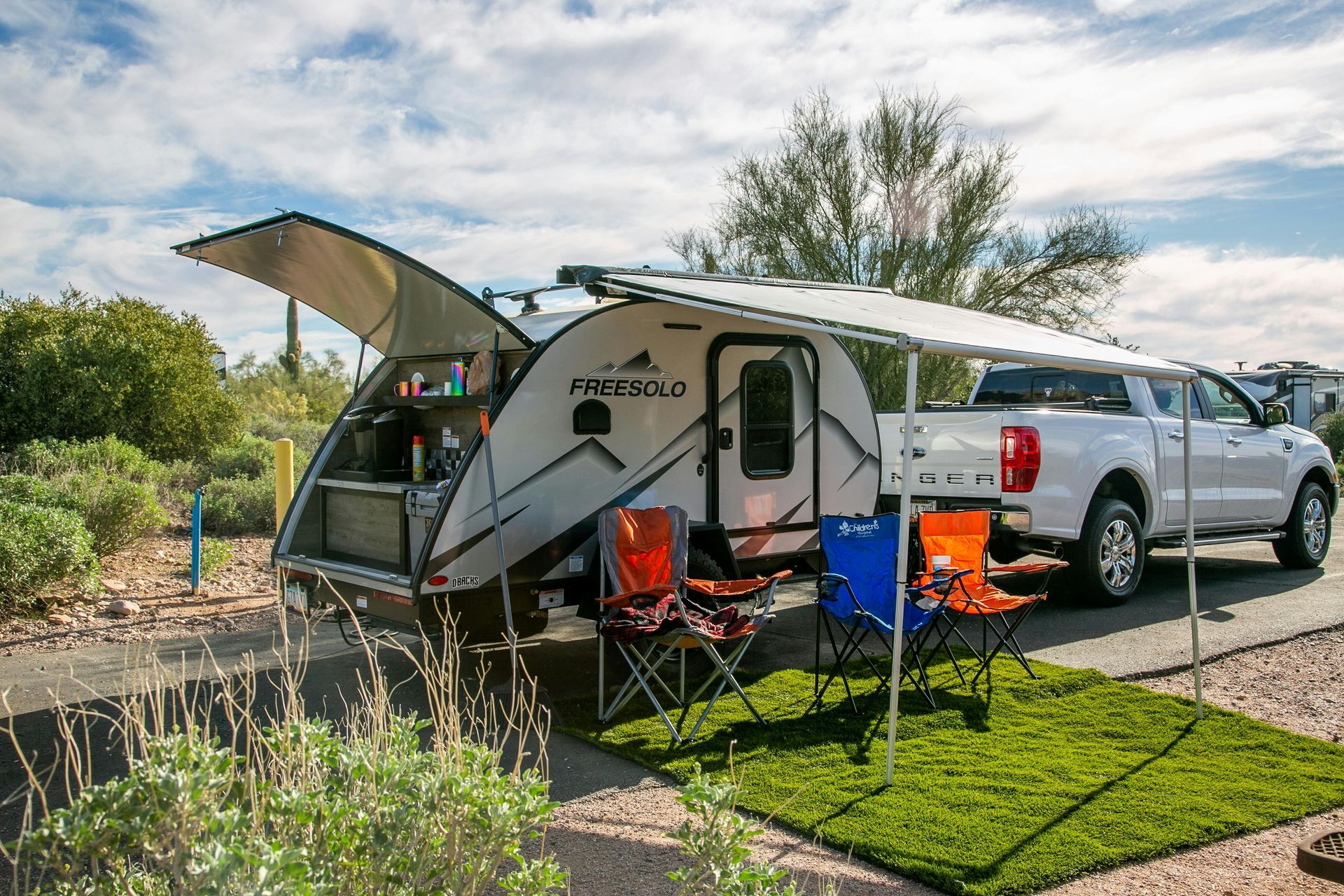 Small camper trailer with awning, chairs, and a pickup truck parked at a campsite.