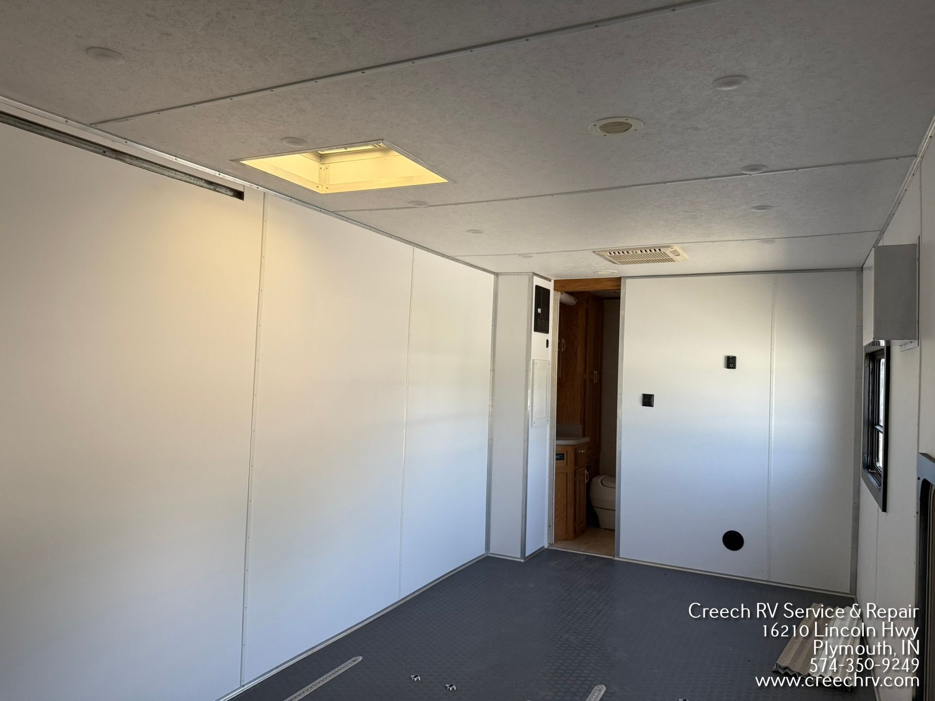 Interior view of an empty RV cargo area with white walls, black floor, and a closed gray rear door.