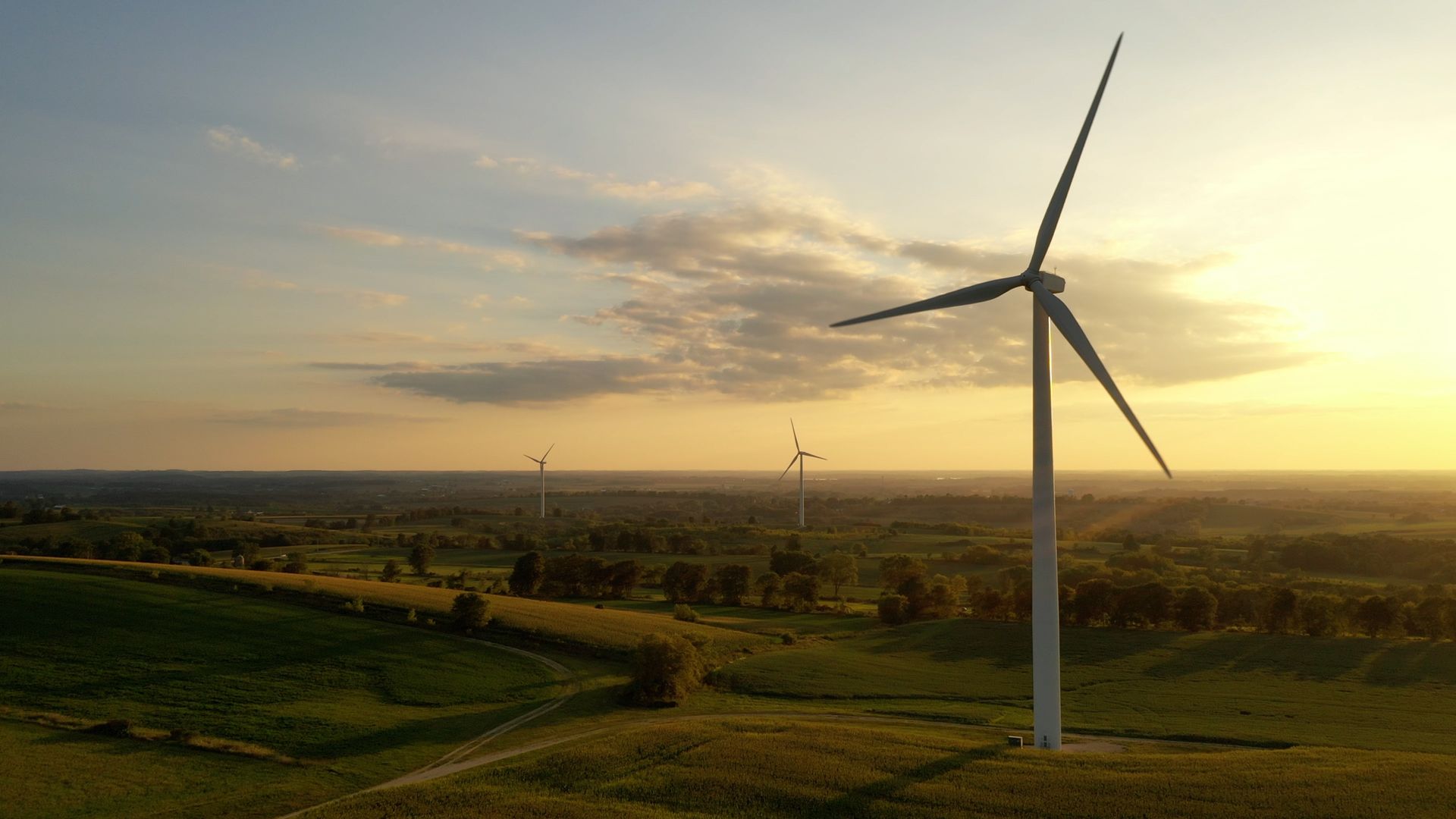 Wind turbine on a grassy hill at sunset, with rolling fields and other turbines in the distance.